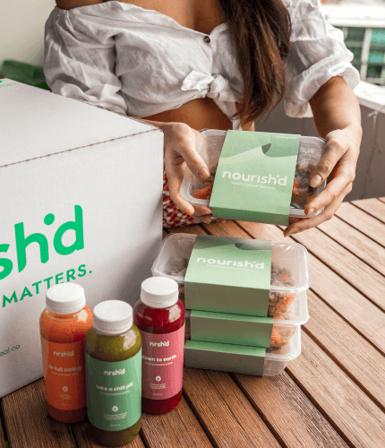 A Nourish'd box on a table, with several meals laid out next to it, in their packaging. A women's hands a shown holding a meal, and there are three bottles of juice on the table