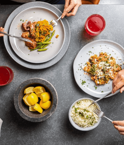 Overhead photo, looking down on a grey marble dining table, laid out with fresh Nourish'd meals, glasses of kombucha, and the hands of people reaching in with cutlery to eat their meal.