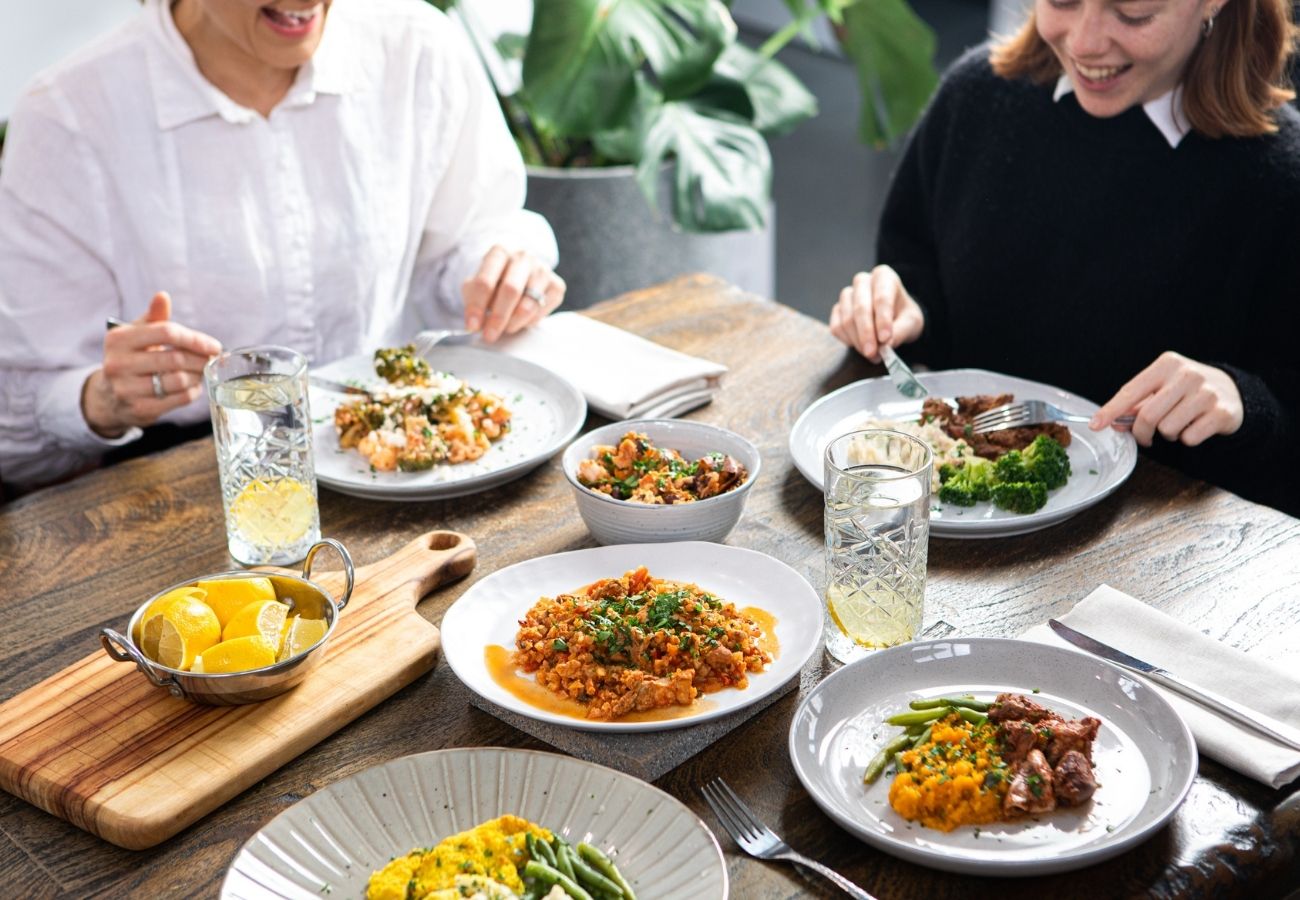 Two young women sitting at a dining table laid out with several Nourish'd meals, about to tuck in to their lunch.