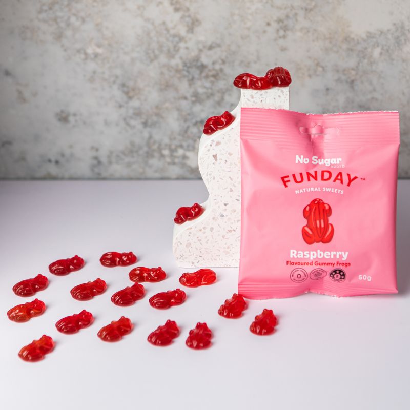 Red gummy frogs placed on a white bench top with a marble background leading to a pink packet leaning against a marble style decorative bookend.