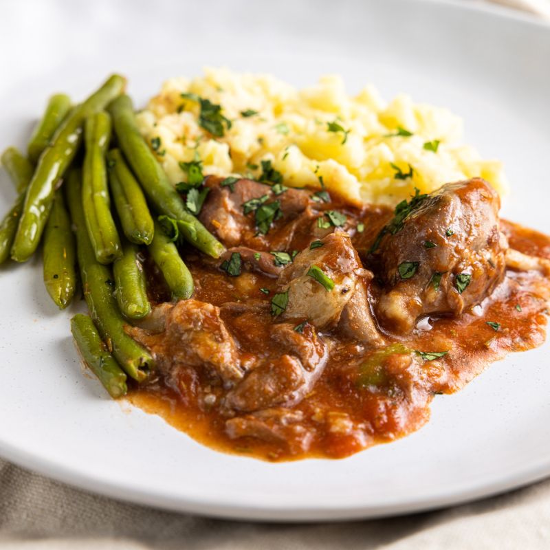 A close up of a rich tomato sauce covered lamb shanks, off the bone, served on a white plate with green beans and mashed potato.