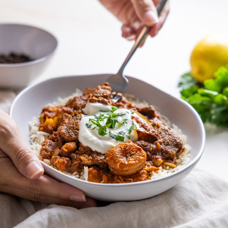 An action shot of Moroccan Beef and Apricot Tagine with Basmati Rice. A hand is holding the bowl while another hand uses a fork to pick up a piece of tender beef. The vibrant dish is garnished with Greek yogurt and spring onion. A ramekin of cracked peppercorns and a lemon are visible in the background, adding fresh elements to the scene.