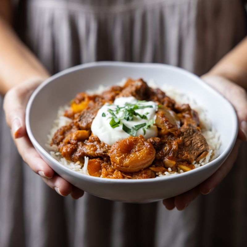 A woman wearing a purple-hued dress holds a white bowl of Moroccan Beef and Apricot Tagine with Basmati Rice. The dish’s rich colors—tender beef, apricots, and pumpkin over basmati rice—stand out against the simple, neutral background.