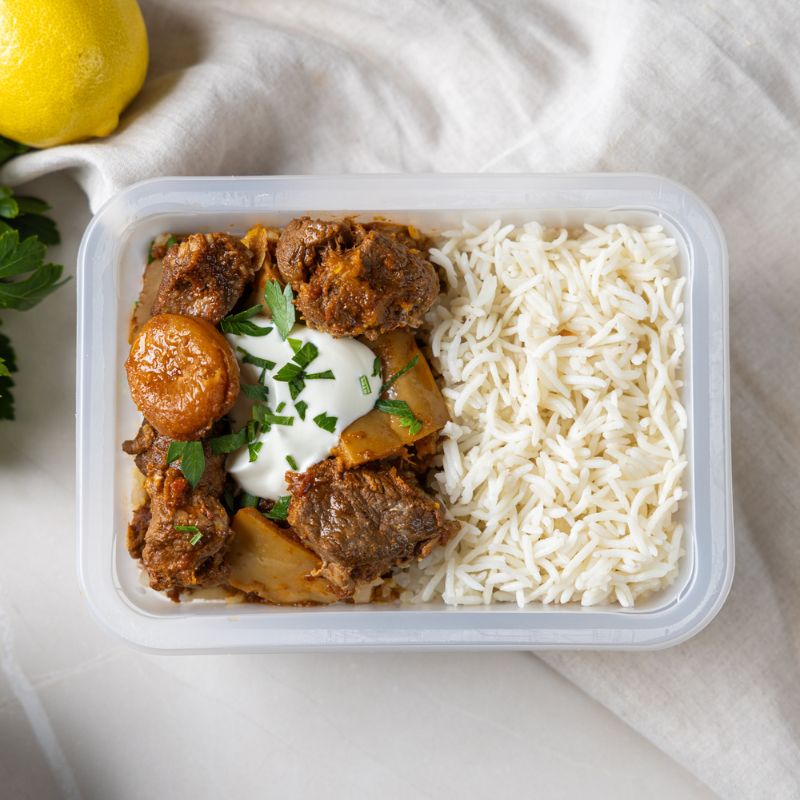 A clear plastic container of Moroccan Beef and Apricot Tagine with Basmati Rice, showcasing the tender beef, apricots, pumpkin, and aromatic spices served on basmati rice. A lemon is visible in the background as a decorative element, adding a fresh and zesty touch to the composition