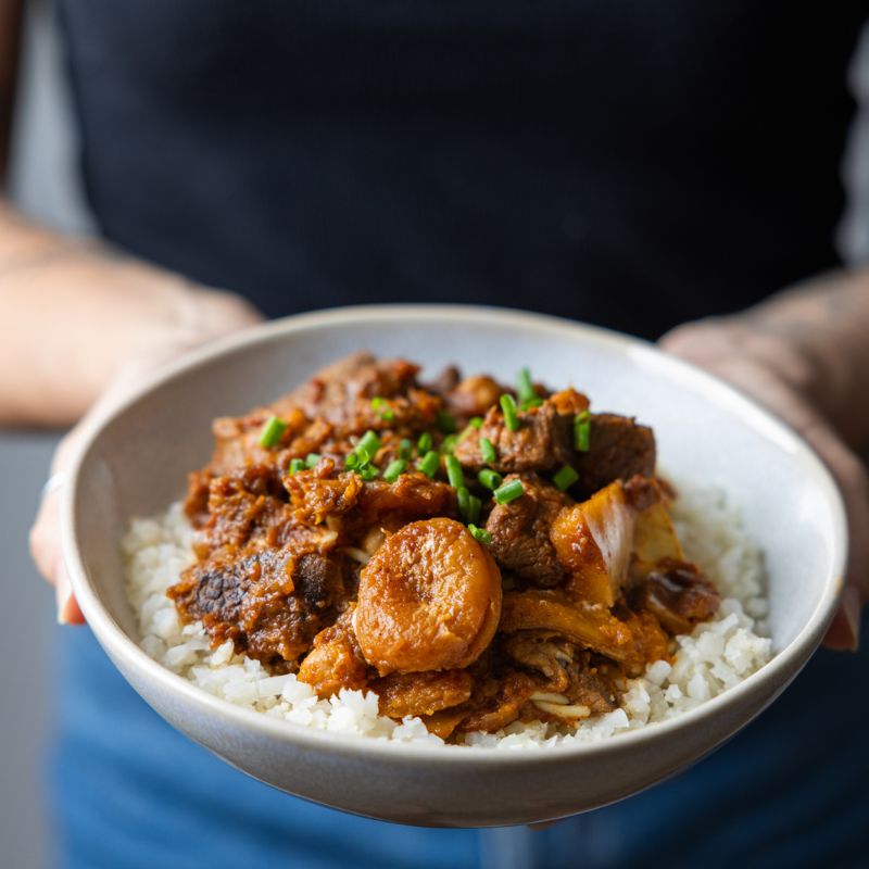 A woman in a black top and blue jean shorts is holding a bowl of Moroccan Beef and Apricot Tagine with Cauliflower Rice. The bowl highlights the vibrant colors of the dish, with beef, dried apricots, and pumpkin sitting atop cauliflower rice. The woman's hands gently cradle the bowl, emphasizing the comforting nature of the meal.