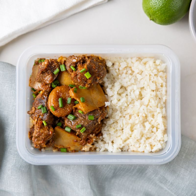 A clear plastic container of Moroccan Beef and Apricot Tagine with Cauliflower Rice, showcasing the hearty beef, dried apricots, pumpkin, and cauliflower rice. A fresh lime is placed in the background as a decorative element, adding a pop of green to the shot.