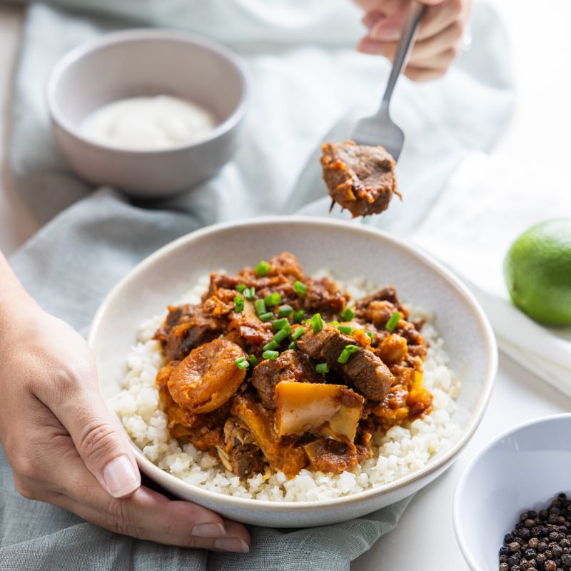 An action shot of Moroccan Beef and Apricot Tagine with Cauliflower Rice. A hand is holding the bowl, while another hand uses a fork to pick up a piece of tender beef. The dish is vibrant with its mix of beef, apricots, and pumpkin. A ramekin of yogurt, cracked peppercorns, and a lime are visible in the background.