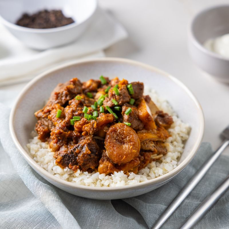 A side-angle view of Moroccan Beef and Apricot Tagine with Cauliflower Rice. The dish is plated in a white bowl, with tender beef, dried apricots, and pumpkin resting on cauliflower rice. The side angle highlights the textures of the meal, with the cauliflower rice and rich sauce adding layers of visual appeal.