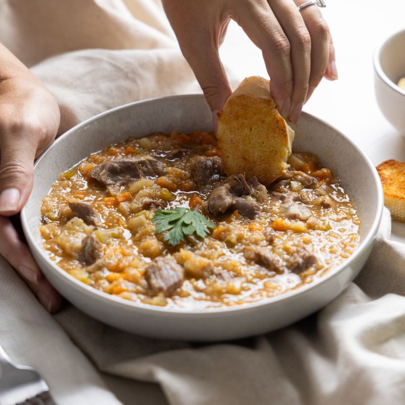 Action shot capturing one hand holding the bowl of Lamb Shank Scotch Broth while the other dips a piece of garlic bread into the broth. The warm, hearty meal is framed by the beige cloth, utensils, and a glimpse of parsley in the corner.