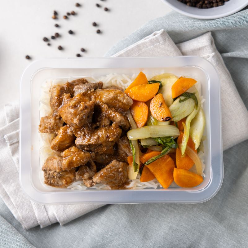 Clear container shot of Sticky Plum Pork, highlighting the vibrant presentation of the pork, rice, and vegetables. Black peppercorns and a white ramekin add texture and detail in the background.