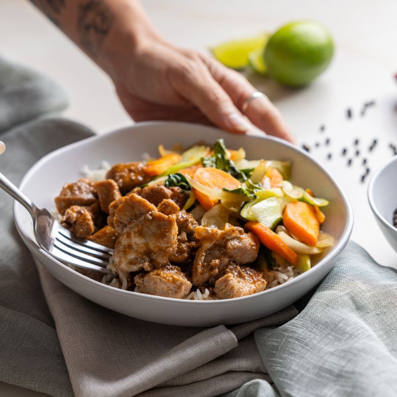 Action shot of two hands; one hand is holding the white bowl of Sticky Plum Pork, while the other is scooping up a portion. A white ramekin of black peppercorns, scattered peppercorns, and a sliced lime can be seen in the background, adding a zesty touch.