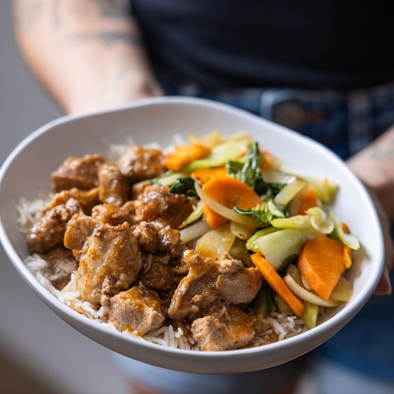 Close-up of Sticky Plum Pork, focusing on the glossy plum sauce coating the pork, vibrant carrot and bok choy, and the fluffy rice base. A person’s arm is visible holding the bowl.