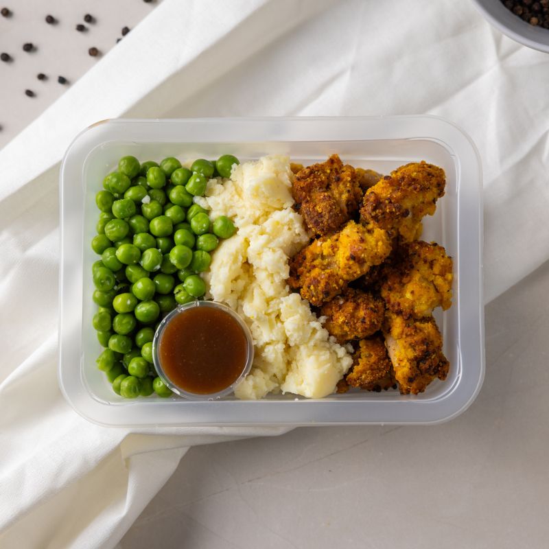 A clear plastic container of Popcorn Chicken with Mash, showcasing crispy popcorn chicken, creamy mashed potatoes, and sweet peas. A small pod of Nourish’d BBQ sauce is included inside the container, with scattered black peppercorns adding a rustic touch to the composition.