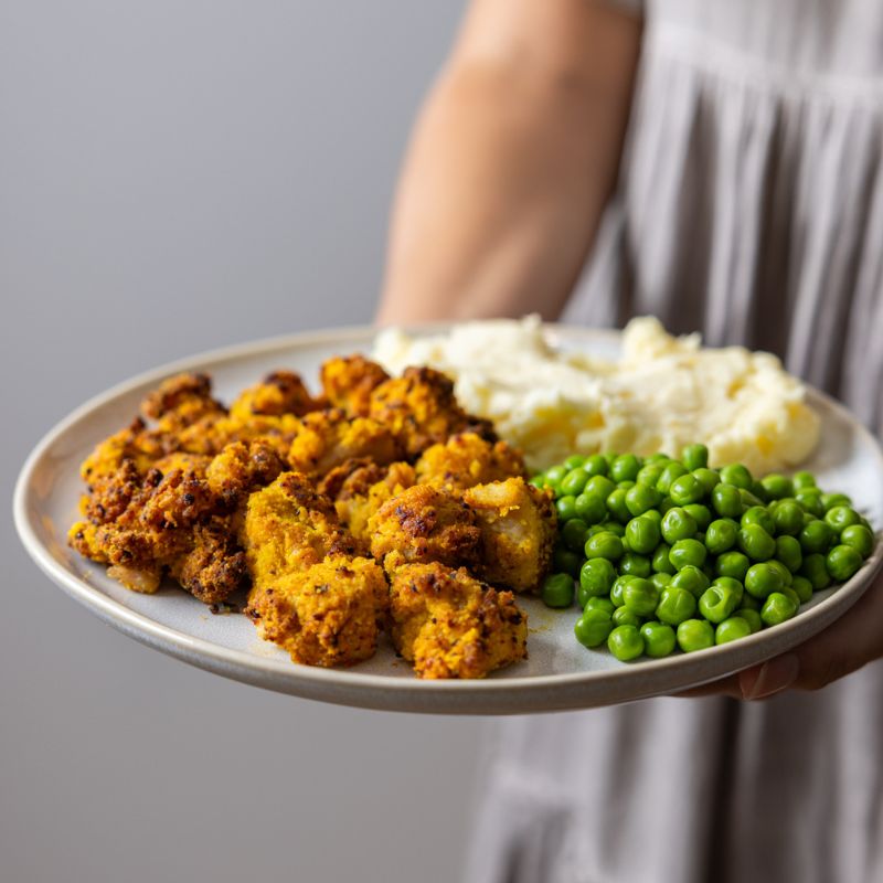 A woman wearing a purple-hued dress holds a white plate of Popcorn Chicken with Mash. The dish features golden popcorn chicken, mashed potatoes, and sweet peas, complemented by a drizzle of tangy BBQ sauce.