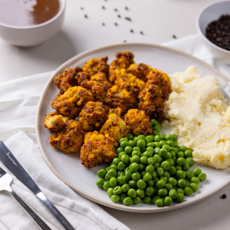 A side angle shot of Popcorn Chicken with Mash on a white plate. The dish includes golden popcorn chicken, creamy mashed potatoes, and sweet peas, paired with a ramekin of cracked peppercorns and a ramekin of gravy. A fork and knife sit beside the plate, with scattered peppercorns adding texture and detail.