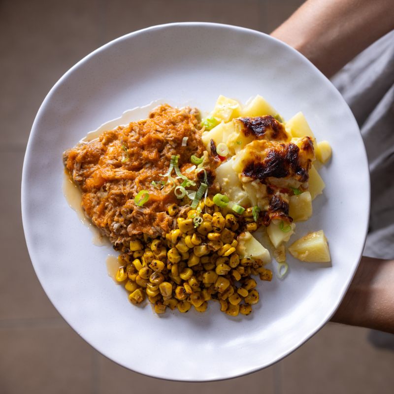 Top view of BBQ Pork Shoulder with Delmonico Potato on a white plate, held by a person’s hands, showcasing the tangy BBQ sauce, creamy Delmonico potatoes, and sweet corn.