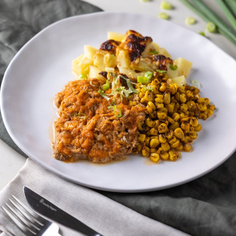  Side shot of BBQ Pork Shoulder with Delmonico Potato, highlighting the layers of tender pork, BBQ sauce, creamy potatoes, and vibrant sweet corn with a spring onion garnish.