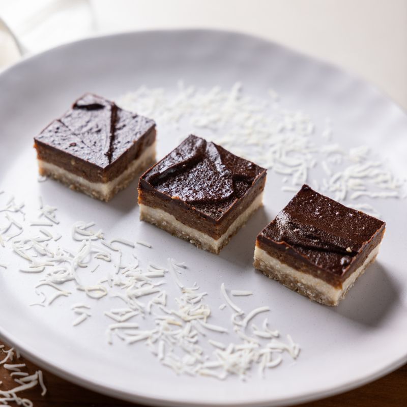 A close-up of three salted caramel slices on a white plate, decorated with dessicated coconut, sitting on a white bench