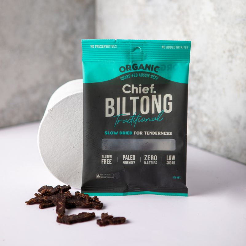 A packet of biltong leaning against a marble prop with some beef biltong pieces in front, all displayed on a white bench with a dark marble backdrop.