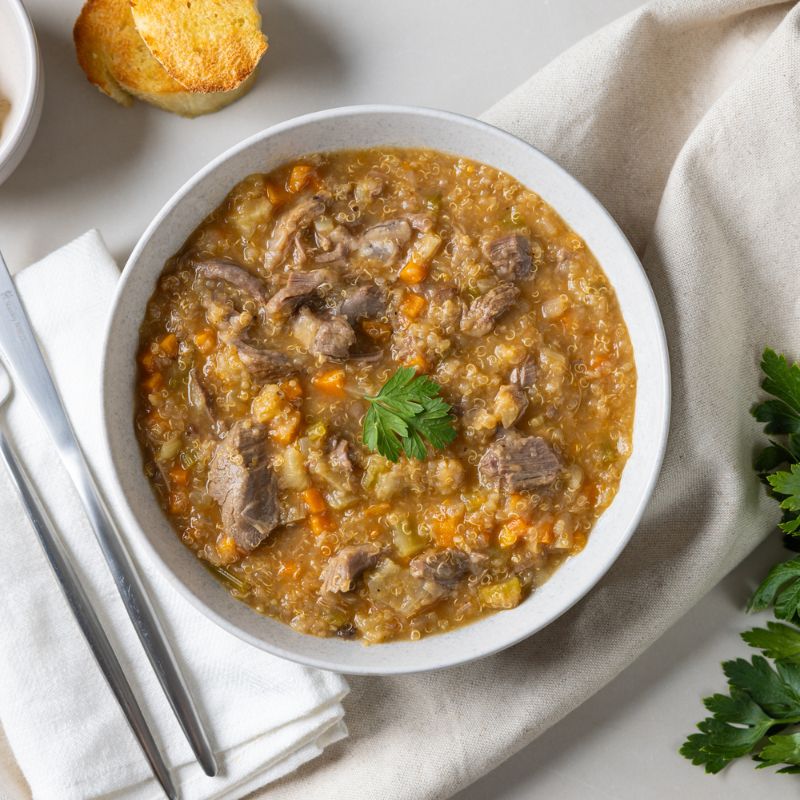 Clear container shot showcasing the Lamb Shank Scotch Broth in all its hearty glory. The container rests on the beige cloth with a fork and knife nearby, adding a simple, practical touch.