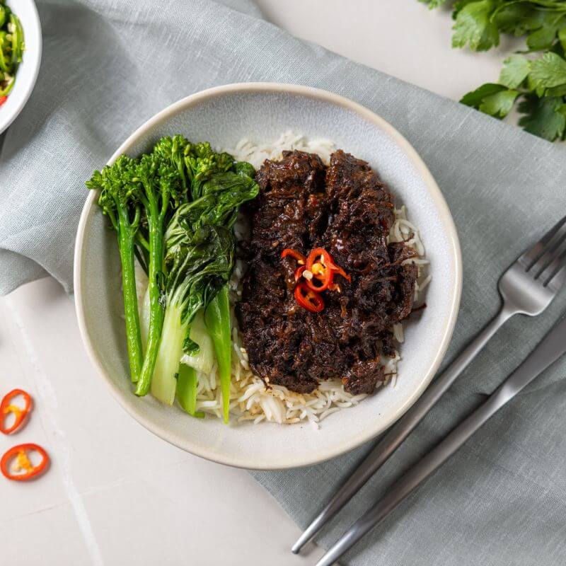 A side-angle shot of Beef Rendang, highlighting the depth of the dish. Tender slow-cooked beef, vibrant bok choy, and broccoli sit atop a bed of basmati rice, with red chili slices providing a pop of color