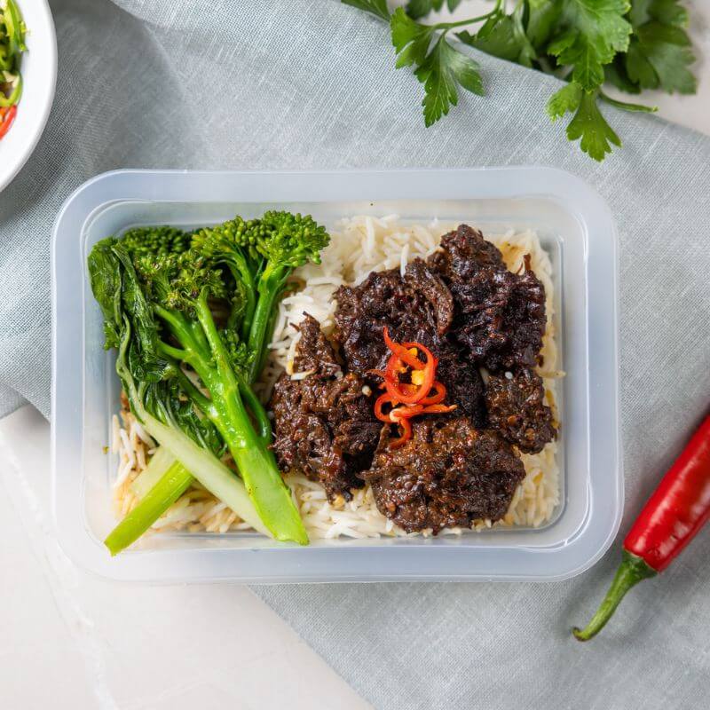 A clear plastic container filled with Beef Rendang, featuring tender beef, bok choy, and broccoli on a bed of basmati rice. The container rests on a light green cloth, with a red chili placed to the right as a decorative element.