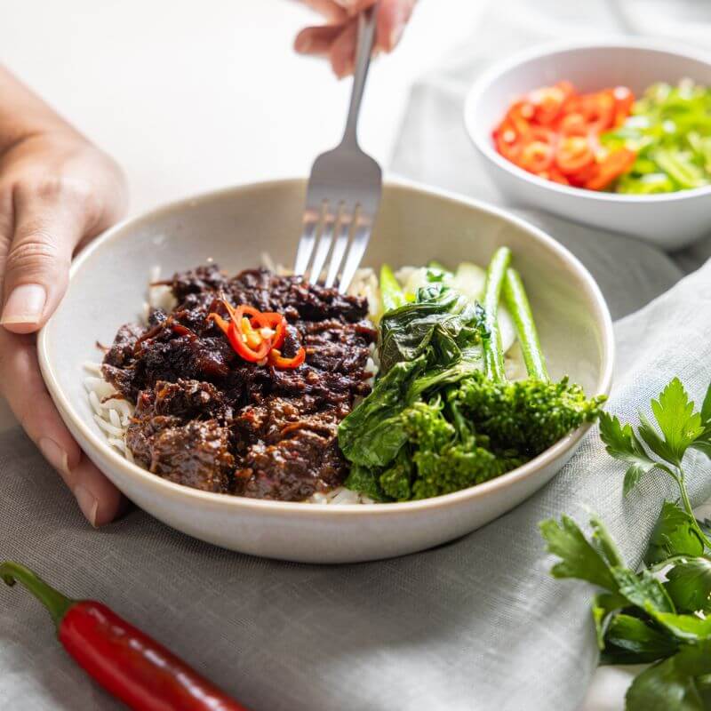 An action shot of Beef Rendang, with one hand holding the white bowl and the other hand using a fork to scoop up a tender piece of beef. The rich, coconut milk sauce and vibrant vegetables are visible