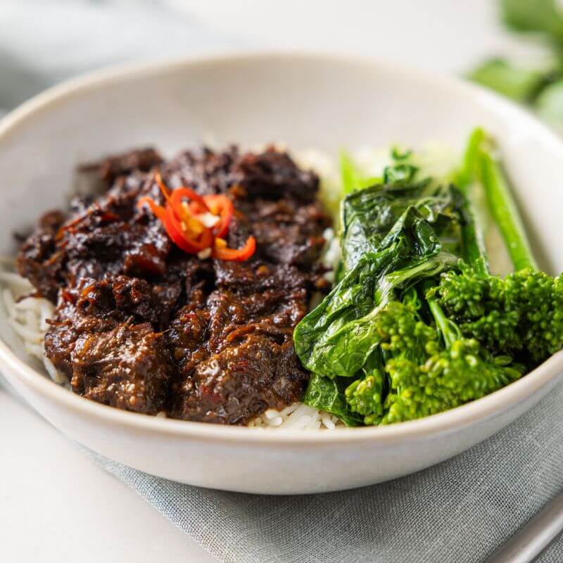 A close-up of Beef Rendang, showcasing the tender slow-cooked beef coated in a rich coconut milk sauce, with bok choy, broccoli, and slices of red chili as garnish.