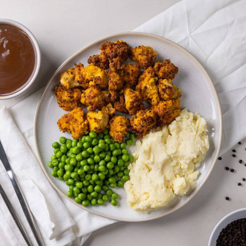 A top-down view of Popcorn Chicken with Mash on a white plate, set on a white counter with a cream-colored cloth underneath. The dish includes crispy, golden popcorn chicken, creamy mashed potatoes, and sweet peas. A ramekin of cracked peppercorns sits in the bottom right corner, with scattered peppercorns and a ramekin of gravy as decorative elements.