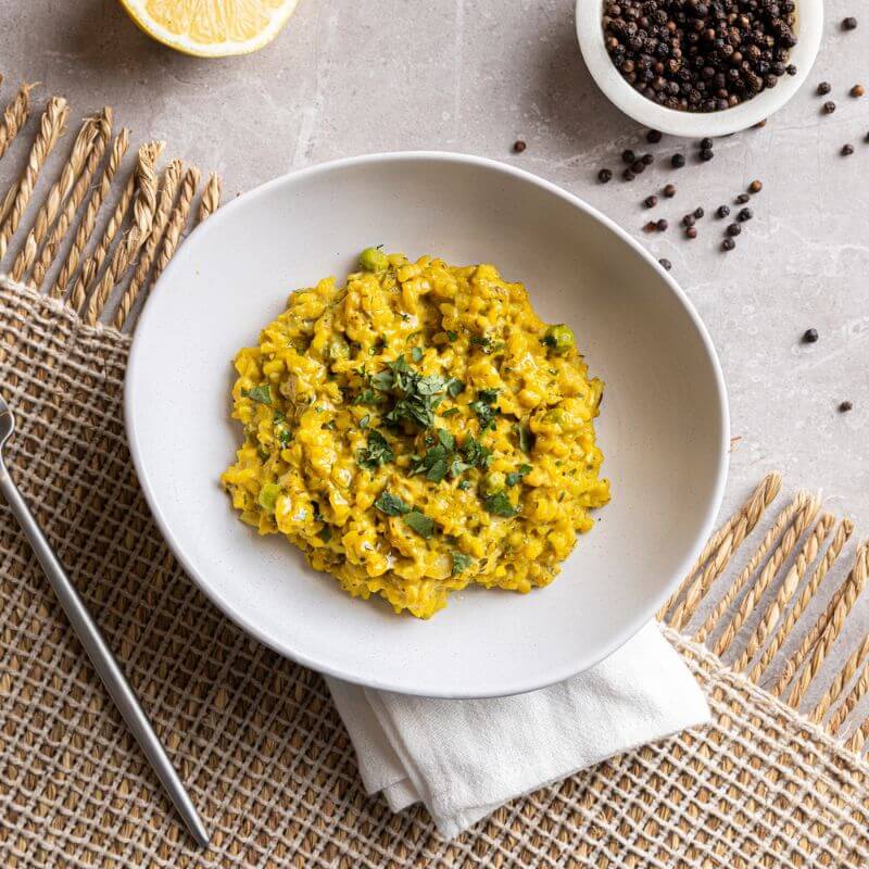 A top-down view of Pumpkin Risotto in a white bowl, set on a wool fringed placemat on a grey marble counter. A cream-colored cloth is tucked underneath the bowl, with a fork to the left. The risotto is garnished with chopped spring onion. Decorative elements include a ramekin of cracked peppercorns in the top right corner, scattered peppercorns, and a sliced lemon.