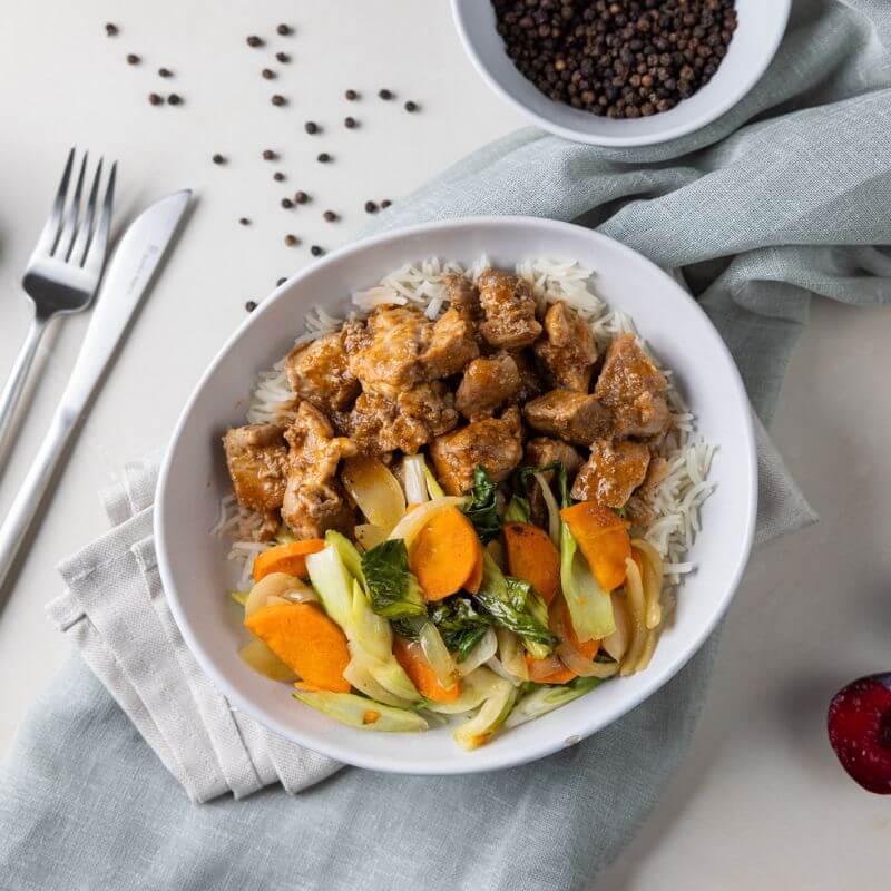 Flat lay shot of Sticky Plum Pork in a white bowl, resting on a beige and grey cloth layered underneath. The dish features tender diced pork in a sweet and tangy plum sauce, served over basmati rice and topped with crisp carrots, tender bok choy, and zesty onions. A fork and knife are placed to the left of the bowl, which is set on a white countertop. A white ramekin of black peppercorns and scattered peppercorns can be seen in the background.