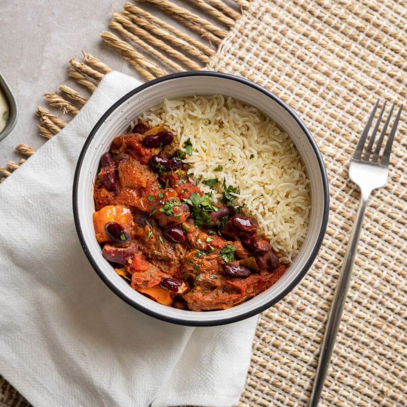 Flat lay shot of Texas Brisket Chilli in a white bowl with a black edge, resting on a rustic tan fringed placemat. A fork is placed to the right of the bowl, and a white napkin is underneath the bowl. The chilli is garnished with parsley for added freshness.