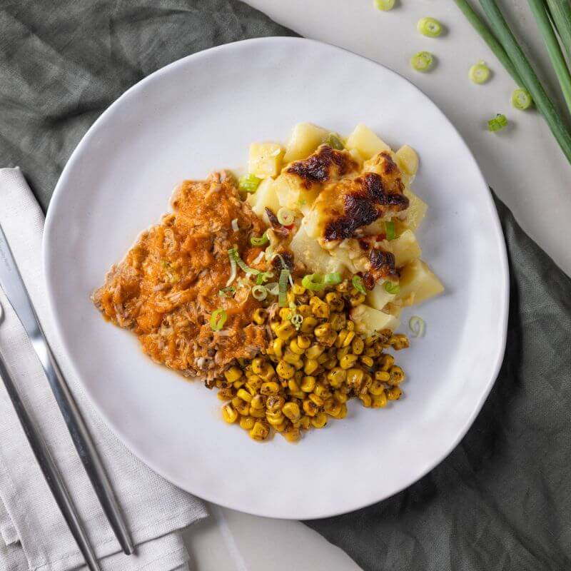 Flat lay shot of BBQ Pork Shoulder with Delmonico Potato on a white plate, placed on a grey cloth. The meal is garnished with chopped spring onion, and a napkin with a knife and fork sits on the left-hand side. Chopped spring onion is visible in the background.