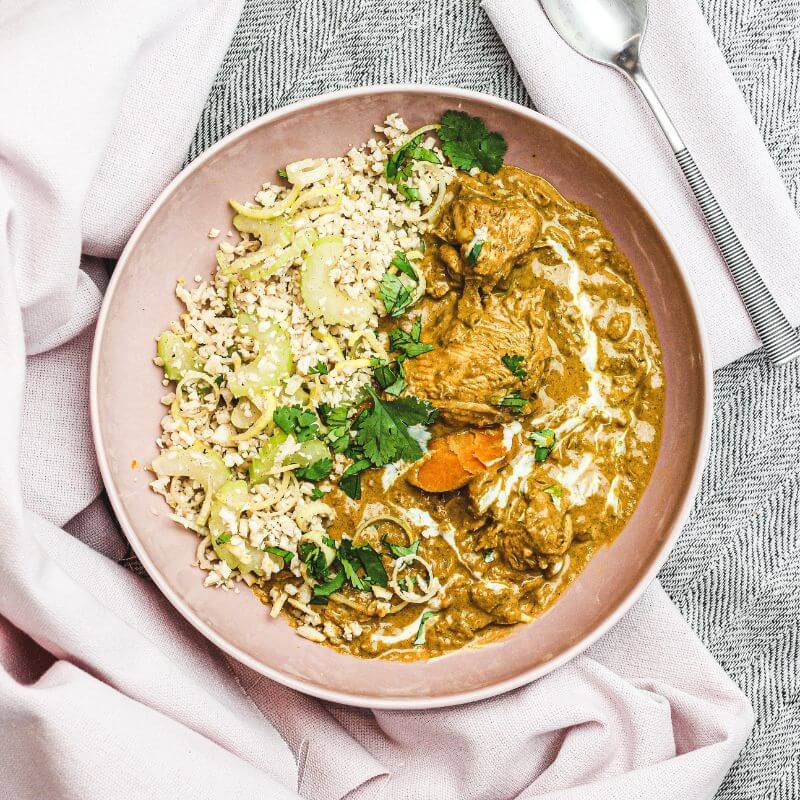 A top-down view of Chicken Masala with Cauliflower Rice served on a light pink plate. The dish features tender chicken in a rich, spiced Indian-style gravy, paired with nutrient-packed cauliflower rice and garnished with chopped parsley. A spoon rests on a white napkin to the right of the plate, while a light pink cloth is tucked underneath. A black and white patterned cloth is placed on the right for added texture, all set against a cozy, inviting backdrop