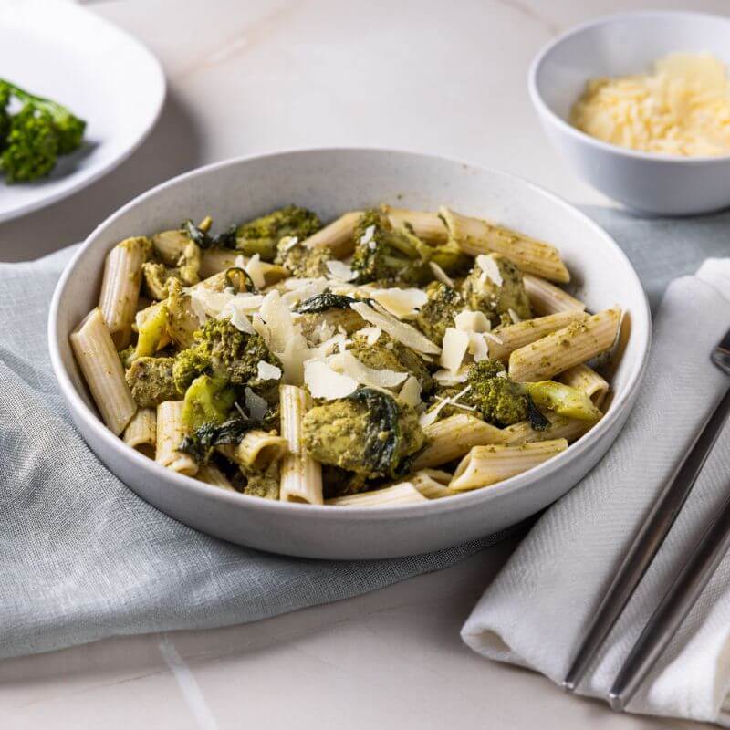 Side view of Chicken Pesto Penne in a white bowl with grated Parmesan cheese, a fork and knife on a napkin, and a plate of broccolini and ramekin of Parmesan cheese in the background.