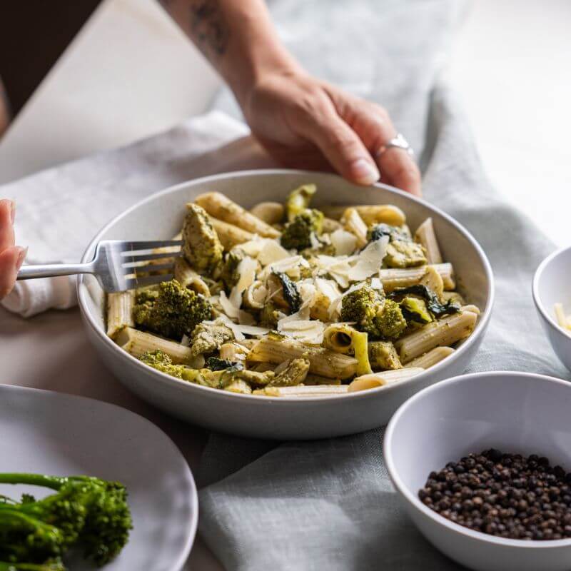 Action shot of a person's hand holding a bowl of Chicken Pesto Penne while using a fork to pick up a piece of chicken, showcasing the tender chicken breast and vibrant pesto sauce.