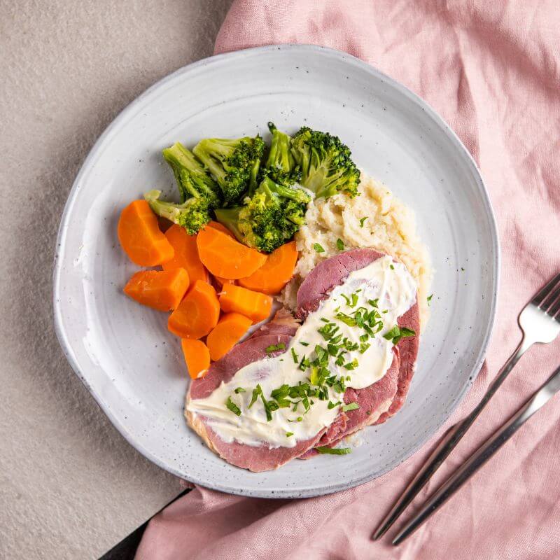 A top-down view of Corned Beef with Cauliflower Mash & White Sauce served on a white plate. The dish features tender slices of slow-cooked silverside, creamy cauliflower mash, steamed vegetables, and a rich white onion sauce, all garnished with chopped parsley. A fork and knife are placed to the right of the plate, and a pink cloth is tucked underneath.