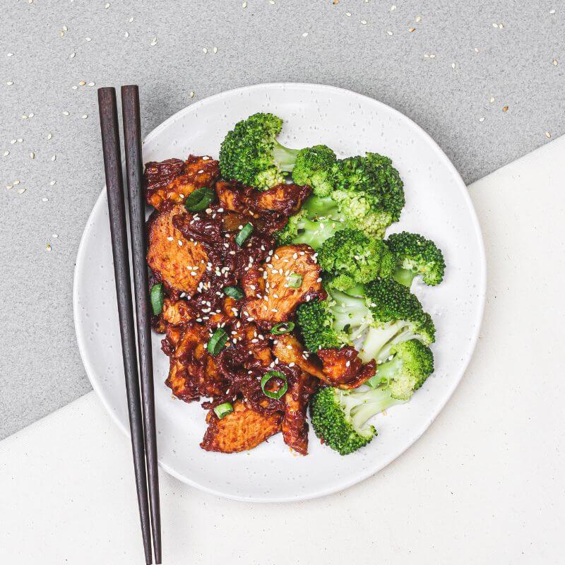 Flat lay shot of Korean Sesame Chicken and Broccoli on a white plate against a marble background, with chopsticks resting on the plate and sesame seeds scattered across the background.