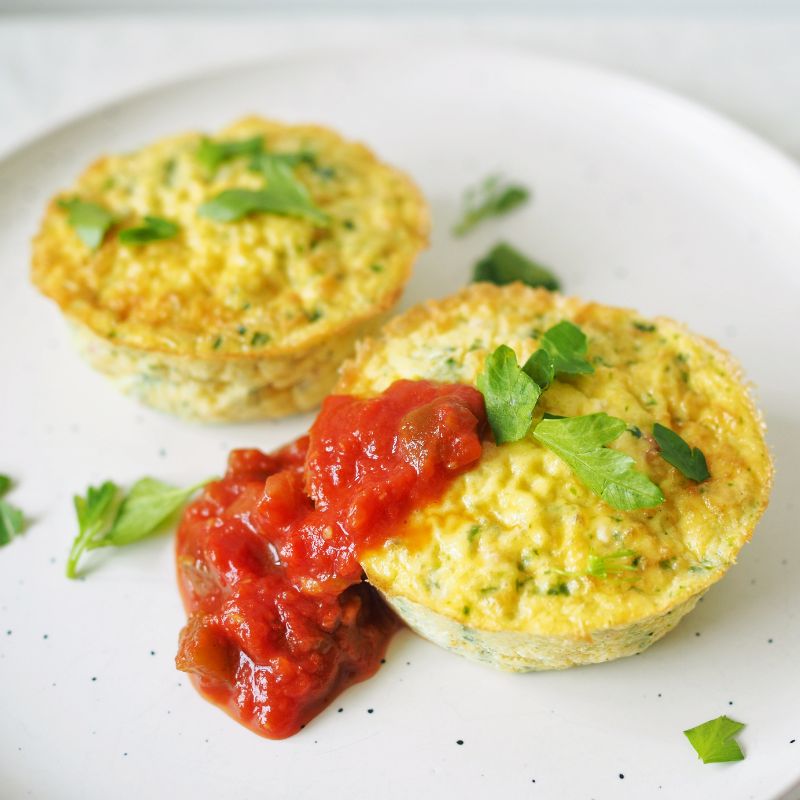 A side angle close-up photograph of a muffin shaped frittata on a white marble plate, scattered with herbs and topped with a tomato salsa.