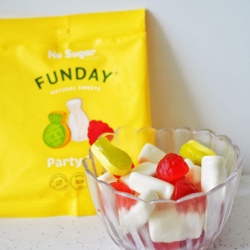 A side on photograph of a small glass bowl containing raspberry, pineapple and milk bottle gummy lollies, served on a white bench top. Behind the bowl is a yellow packet the gummies are delivered in leaning against a white back drop.