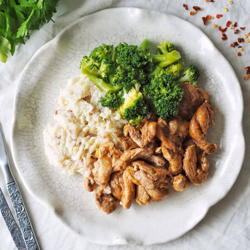 Flat lay image of Portuguese Chicken served on a white curved plate. The dish includes tender strips of RSPCA-approved chicken coated in Portuguese spices, creamy cauliflower mash with bacon, and broccoli. The plate sits on a white cloth, with a fork and knife to the left. Garnishes of parsley and scattered chili flakes add a vibrant touch in the background.