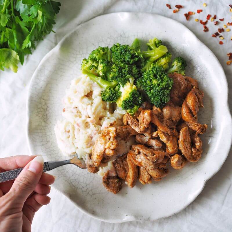 Action shot of a hand holding a fork, picking up a piece of Portuguese Chicken from the plate. The setup includes the same elements as above: white curved plate, white cloth, parsley garnish, and chili flakes scattered for decoration.