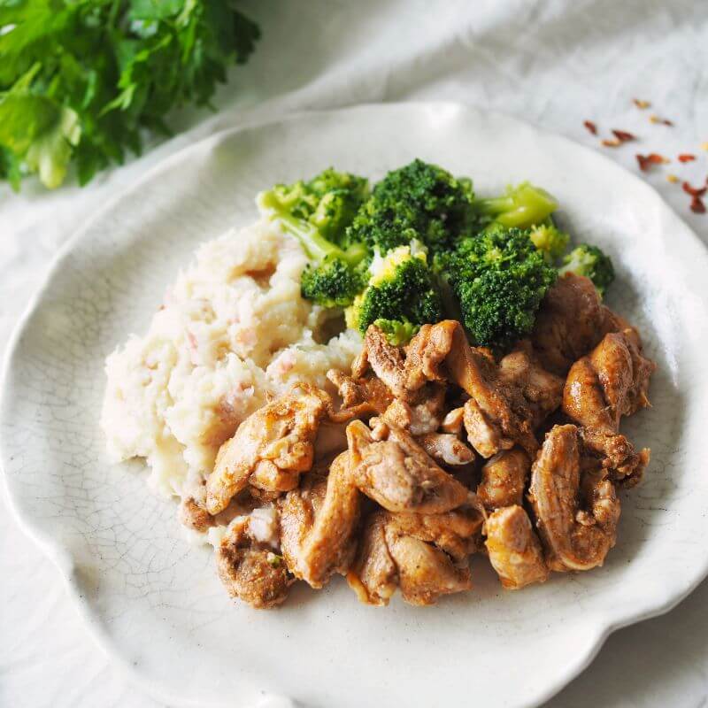 Side view of the Portuguese Chicken plate, highlighting the creamy cauliflower mash and tender chicken strips. The background elements remain consistent, with parsley garnish and scattered chili flakes.