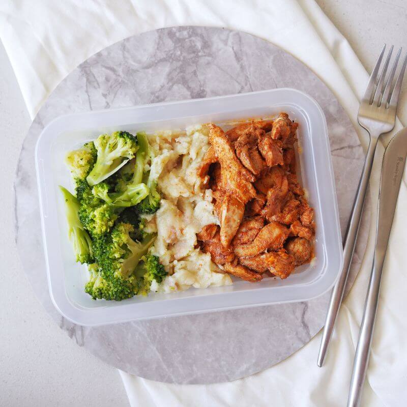 Clear container shot of Portuguese Chicken, showing its convenience for portability. A knife and fork are placed to the right of the container, which is set on a marble placemat over a white cloth for a clean and modern presentation.