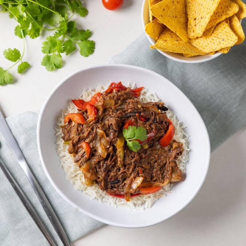 A top-down view of Beef Fajitas served in a white bowl placed on a light green cloth. The dish features tender strips of grass-fed beef, capsicum, and onions, served on a bed of basmati rice. A fork and knife sit to the left of the bowl, with a bunch of fresh cilantro in the top left corner and a single cherry tomato near the top. A white ramekin filled with corn chips adds a touch of Mexican flair. The setup rests on a clean white countertop.