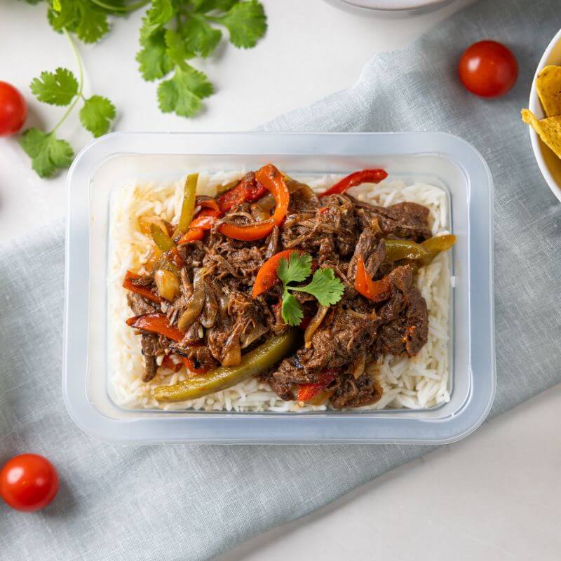 A clear plastic container of Beef Fajitas resting on a light green cloth over a white countertop. The container showcases strips of tender beef, capsicum, and onions on basmati rice, garnished with scattered cherry tomatoes. A bunch of fresh cilantro and a ramekin of corn chips are visible in the background, adding vibrant and authentic elements to the composition.