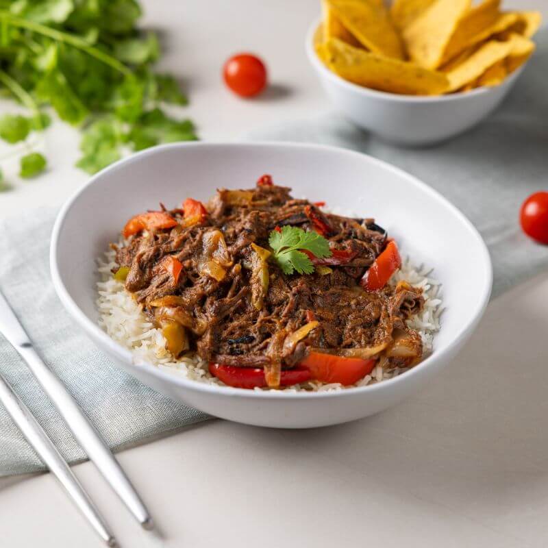 A side-angle shot of Beef Fajitas served in a white bowl, resting on a light green cloth atop a white countertop. The dish features tender beef strips, capsicum, and onions on a bed of basmati rice. A bunch of cilantro, scattered cherry tomatoes, and a ramekin of corn chips complement the scene, creating a visually rich presentation.