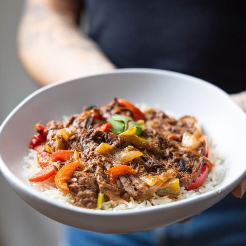 A close-up shot of a woman holding a white bowl of Beef Fajitas with both hands. The dish highlights tender beef strips, capsicum, and onions, served over basmati rice. The woman is wearing a black top and blue jean shorts, adding a casual and relatable touch to the image.