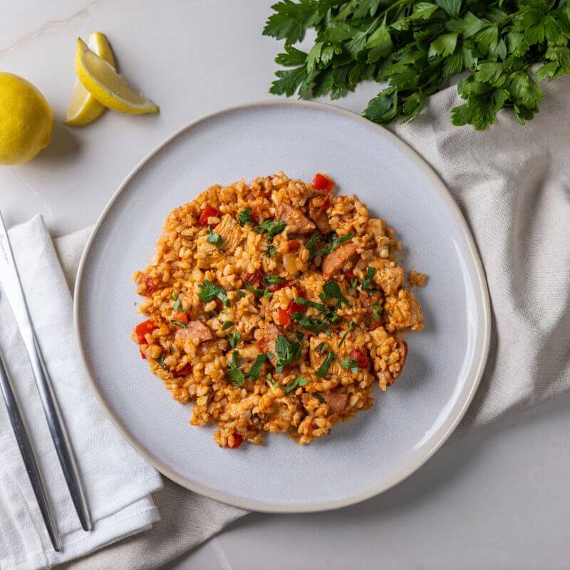 A top-down view of Paella with Brown Rice served on a white plate. The dish features tender chicken breast, smoky chorizo, capsicum, onion, and tomato, all served on a bed of brown rice and garnished with chopped cilantro. The plate is placed on a white cloth on a clean white countertop. A bunch of fresh cilantro sits in the top right corner, while sliced lemons add a zesty touch to the top left corner. A fork and knife rest to the left of the plate.