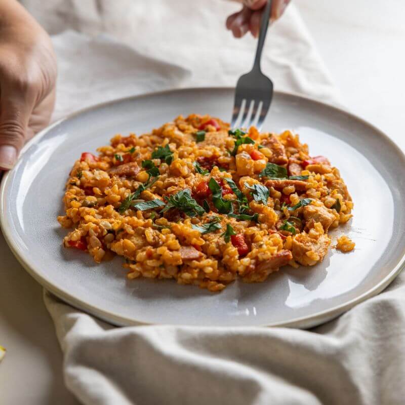 An action shot of Paella with Brown Rice. One hand holds the plate while another hand scoops up a piece of the dish with a fork. The paella features chicken breast, chorizo, capsicum, onion, and tomato on brown rice, garnished with fresh chopped cilantro.